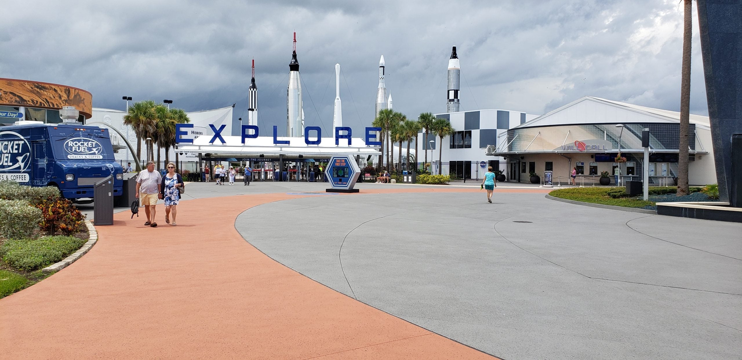 People walk around commercial walkway with rocket ships in the background
